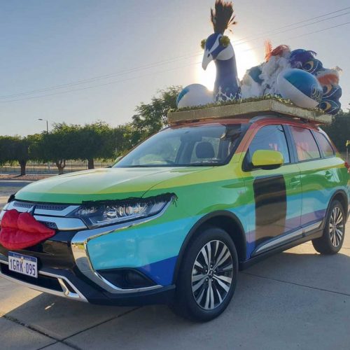 SUV vehicle parked in driveway decorated with a rainbow themed vinyl wrap and a peacock on the roof